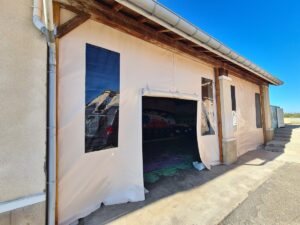 Façade d'un bâtiment avec une grande bâche beige provisoire, comprenant fenêtres transparentes et une ouverture centrale, sous un ciel bleu clair.