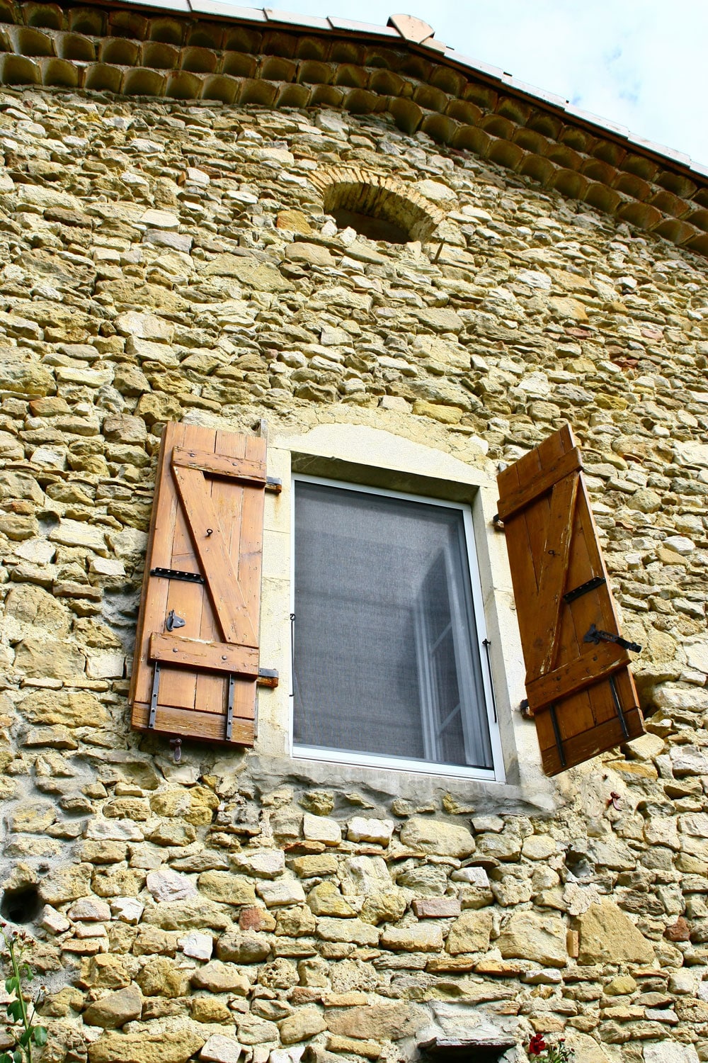 Façade ancienne en pierres jaunes et beige avec une fenêtre moderne à volets bois ouverts, sous une petite lucarne cintrée.